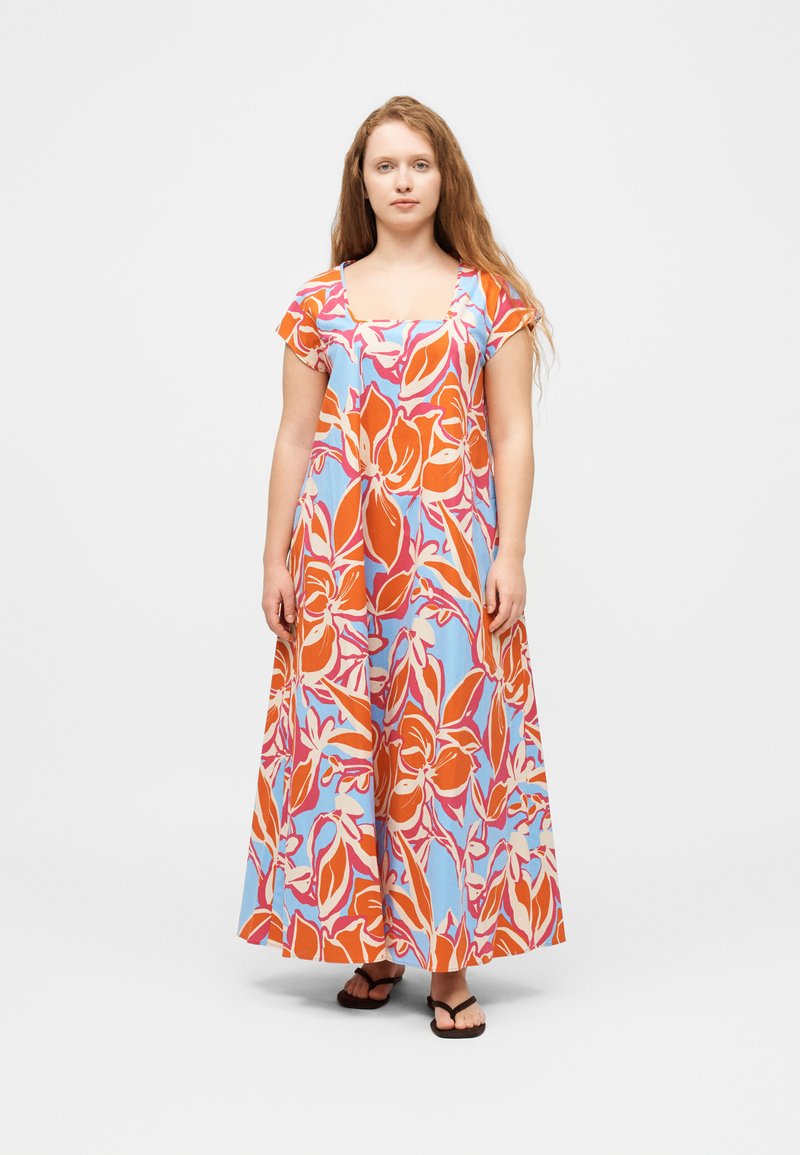 Γυναίκα με μακριά μαλλιά, που φοράει ένα μακρύ φόρεμα με κοντά μανίκια και μεγάλα πορτοκαλί και λευκά λουλουδένια σχέδια σε μπλε φόντο, όρθια.