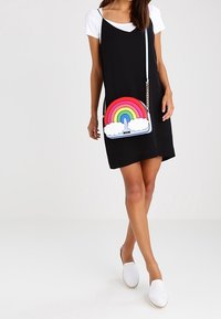 Nainen, jolla on päällään musta liukuväri-mekko valkoisen t-paidan päällä, valkoiset liukuhalvarit ja kantaa värikästä sateenkaaren väristä olkalaukkua, jossa on pilviä.