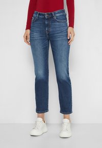 Jeans de mezclilla azul oscuro con corte recto, cintura de media altura y un ligero desvanecimiento. Combinados con zapatillas blancas y una camiseta de manga larga roja.