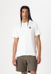 Hvit bomull t-skjorte med et lite brodert motiv på brystet, sammen med olivengrønne cargo shorts med flere lommer.