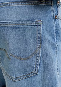 Πίσω τσέπη από ανοιχτόχρωμα τζιν με πορτοκαλί ραφές και μικρή καμπυλωτή λεπτομέρεια ραφής στην τσέπη.