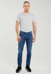 Noor mees seisab, kandes helehallit T-särki, siniseid teksapükse ja musti tossusid, taustaks lihtsalt hele taust.