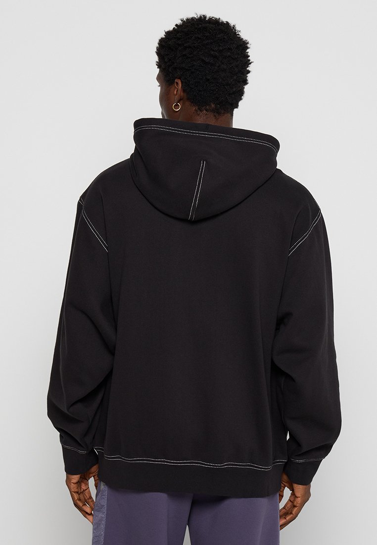Sudadera con capucha negra de corte holgado, con costuras blancas contrastantes y un bolsillo tipo canguro. Tela suave, puños y dobladillo de canalé.