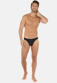 Czarne majtki bikini w kropki, wykonane z elastycznego materiału, z niskim stanem i miękką teksturą, noszone przez męskiego modela.
