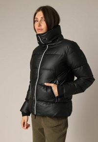 Zwarte gewatteerde jas met hoge kraag, ritssluiting en zijzakken. Heeft een doorgestikt patroon en geribbelde manchetten voor isolatie.