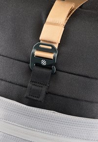 Tessuto nero testurizzato con una sezione in rete grigia chiara. Presenta una tracolla color sabbia e una fibbia in plastica verde scuro con un logo, evidenziando elementi di design funzionali.
