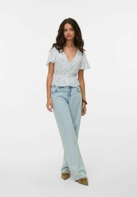 Blusa bianca a fiori con maniche corte a sbuffo e scollatura a V, abbinata a jeans a gamba larga blu chiaro e scarpe ballerine marroni.