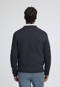 Dunkelgrauer Strickpullover mit geripptem Kragen und Bündchen, mit strukturierter Oberfläche und legerer Passform, getragen über einem hellen Hemd.