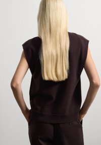 Dunkelbrauner ärmelloser Pullover mit einem gerippten Saum, weicher Textur und strukturiertem Schulterdesign. Langes, glattes, blondes Haar fällt den Rücken hinunter.