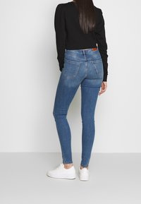 Jeans ajustados de mezclilla en azul medio con una textura suave, cintura alta y diseño de cinco bolsillos. Combinados con zapatillas blancas.