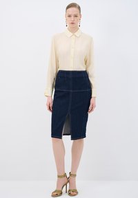 Camisa de botón color amarillo claro, combinada con una falda lápiz de mezclilla oscura con una abertura delantera y costuras blancas en contraste. Tacones verdes con tiras.
