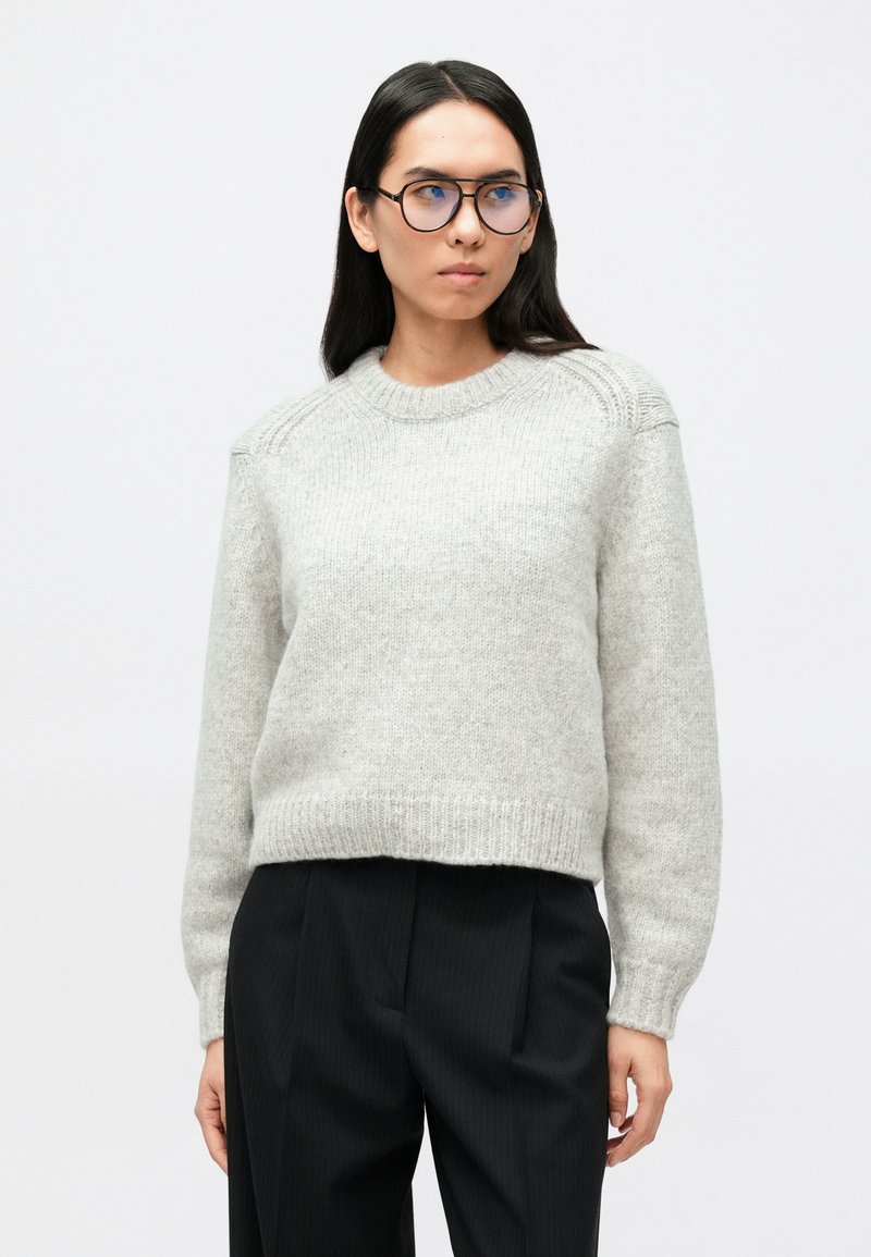 Svetlosivý pletený sveter s okrúhlym výstrihom, rebrovaným lemom a zložitými detailmi na pleciach, v kombinácii s čiernymi nohavicami s prúžkami.