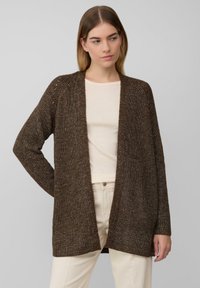 Braune Strickjacke mit offener Vorderseite, langen Ärmeln und einem strukturierten Design. Getragen über einem hellen Oberteil und hellen Hosen.
