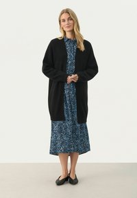 Cardigan negru din tricot purtat peste o rochie midi cu model floral albastru. Cardiganul are mâneci lungi și un design deschis în față. Pantofi negri, cu talpă joasă.