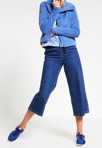 Giacca in pile azzurro chiaro con colletto alto, abbinata a pantaloni in denim blu scuro a girovita e sneakers blu sportive. Texture lisce in tutto.