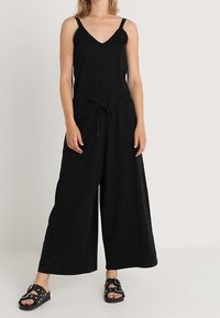 Zwarte jumpsuit met een V-hals, dunne schouderbanden, wijde pijpen, striksluiting in de taille, gemaakt van zachte stof, gecombineerd met zwarte sandalen.