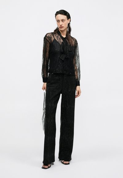 Sort transparent bluse i blonder med lange ærmer, en bindebåndsudskæring, parret med sorte glitrende brede bukser og flade sandaler, mens der bæres en sølvfarvet håndtaske.