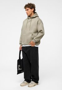 Beige oversized Hoodie mit einer Fronttasche und Kordeln, kombiniert mit schwarzer, weit geschnittenen Hose und beigen Sneakern, während er eine schwarze Tragetasche hält.