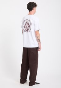 Homme aux cheveux courts et foncés portant un t-shirt blanc avec un motif graphique et un pantalon large marron, de dos sur fond blanc.