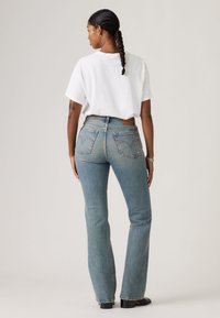 Γυναίκα με πλεξούδες, γυρισμένη πίσω, φοράει λευκό μπλουζάκι, μπλε τζιν ψηλόμεσο, μαύρα παπούτσια και ασημένια βραχιόλια.