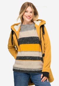 Giacca esterna gialla con cappuccio, indossata sopra un maglione a righe grigio, beige e giallo. Texture accogliente con polsini a coste.