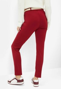 Red stretch trousers with a tapered design, featuring a waistband and a smooth texture, paired with white and burgundy sneakers.