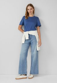 T-shirt bleu, coupe décontractée, associé à un jean large clair. Un sweatshirt blanc noué à la taille et des baskets beiges achèvent le look.