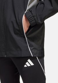 Zwarte sportjack met witte en grijze accenten, voorzien van een elastische tailleband en ritszakken. Zwarte leggings met wit logo.