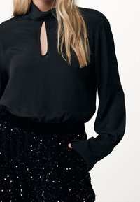 Blusa nera con colletto alto e apertura a chiave; maniche lunghe con polsini arricciati. Abbinata a una gonna nera con paillettes e cintura elastica.