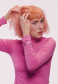 Personne avec des cheveux courts orange et une frange, portant un haut à manches longues rose, ajustant ses cheveux avec des mains ornées de plusieurs bagues en argent et d'un bracelet.