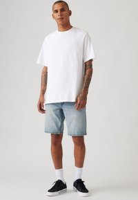 Tricou alb din bumbac cu mâneci scurte, asortat cu pantaloni scurți din denim albastru deschis. Adidași negri și șosete albe completează ținuta.