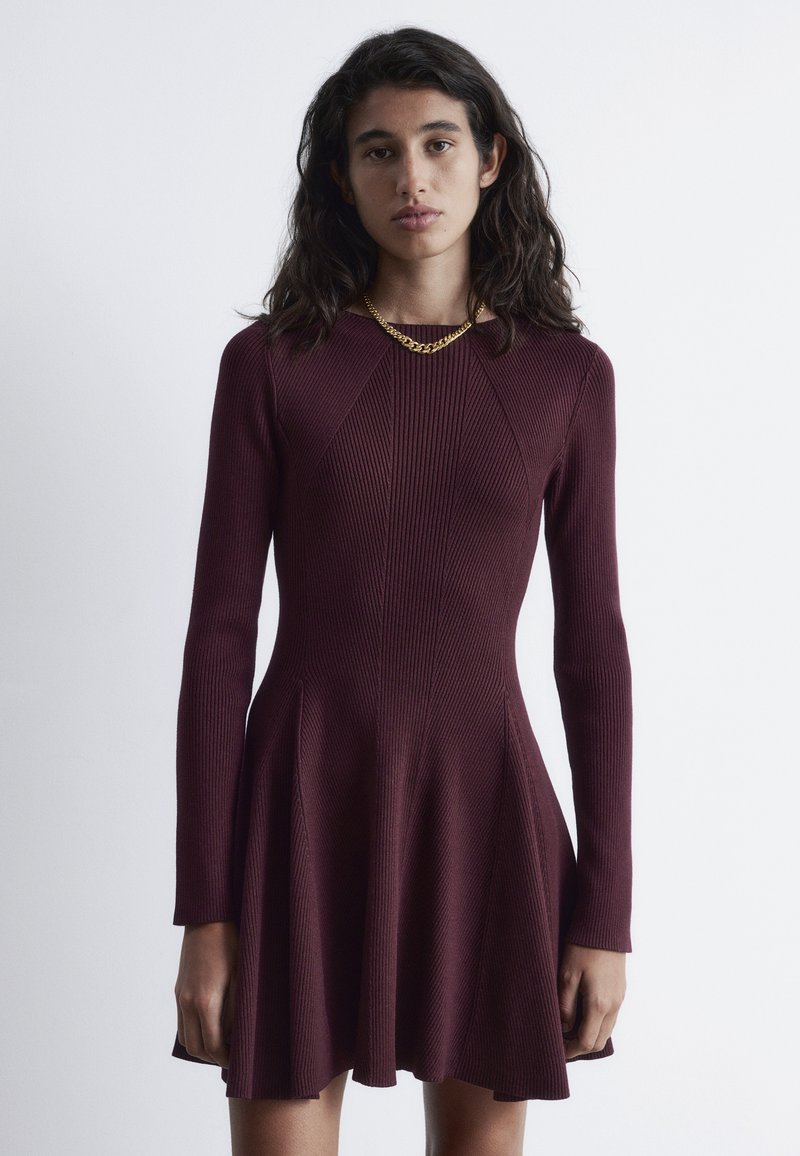 Kastanienbraunes geripptes Kleid mit langen Ärmeln, tailliertem Oberteil, ausgestelltem Rock und geometrischen Nahtdetails. Strukturierter Stoff mit leichtem Glanz.