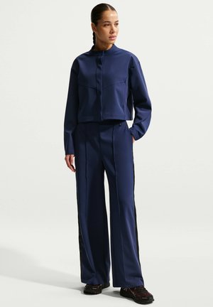 Femme portant une veste zippée à manches longues bleu marine et un pantalon large assorti avec des bandes noires sur les côtés, debout avec une main dans la poche.