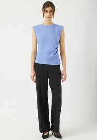 Ljusblå ärmlös topp med rund halsringning, med detaljer i drapering, tillsammans med svarta vida byxor och svarta spetsiga skor.