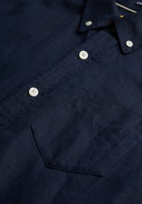 Camisa de botones azul marino hecha de tejido entrelazado. Presenta un bolsillo en el pecho con un logotipo sutil, botones blancos y un cuello.