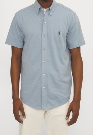 Homme portant une chemise bleu clair à manches courtes avec un petit logo brodé sur la poitrine et un pantalon blanc.