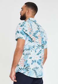 La camicia a maniche corte presenta motivi floreali con foglie tropicali blu e rosa su uno sfondo chiaro, con una vestibilità rilassata e colletto button-down.