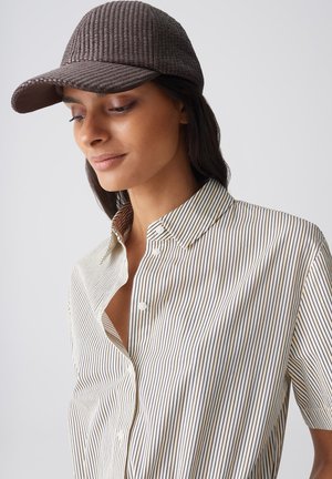 Woman wearing brown textured baseball cap and white short-sleeve shirt with thin vertical black stripes, looking downward with slight smile.