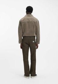 Man met kort krullend haar, gekleed in een taupekleurig jack, donkergroene broek met achterzakken, en witte sneakers met blauwe details, staand met de rug naar de kijker.