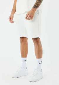 Heren witte shorts van textuurstof, met zijzakken. Draagt witte sneakers en enkelsokkens van wit met zwart logo.