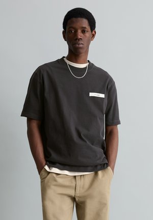 Mladý muž oblečený v tmavom nadrozmernom tričku, béžových nohaviciach a striebornej retiazke, stojaci s rukami v rukávoch na jednoliatom sivom pozadí.