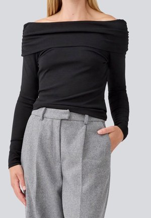 Femme portant un haut noir à épaules dénudées avec manches longues et un pantalon gris à motif chevrons taille haute, une main dans la poche.