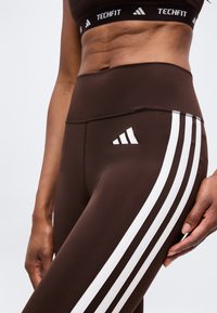 Personne en forme portant des leggings Adidas Techfit marron avec des bandes blanches et un soutien-gorge de sport assorti, debout avec la main détendue le long du corps.