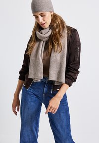 Un bonnet en tricot beige et une écharpe assortie drapés autour du cou, superposés sur un haut marron et une veste bomber marron foncé, associés à un jean bleu.