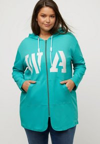 Chaqueta con capucha en color turquesa, con cremallera y texto en blanco, que presenta bolsillos frontales y una textura suave. Las mangas son largas y cuentan con lazos ajustables.