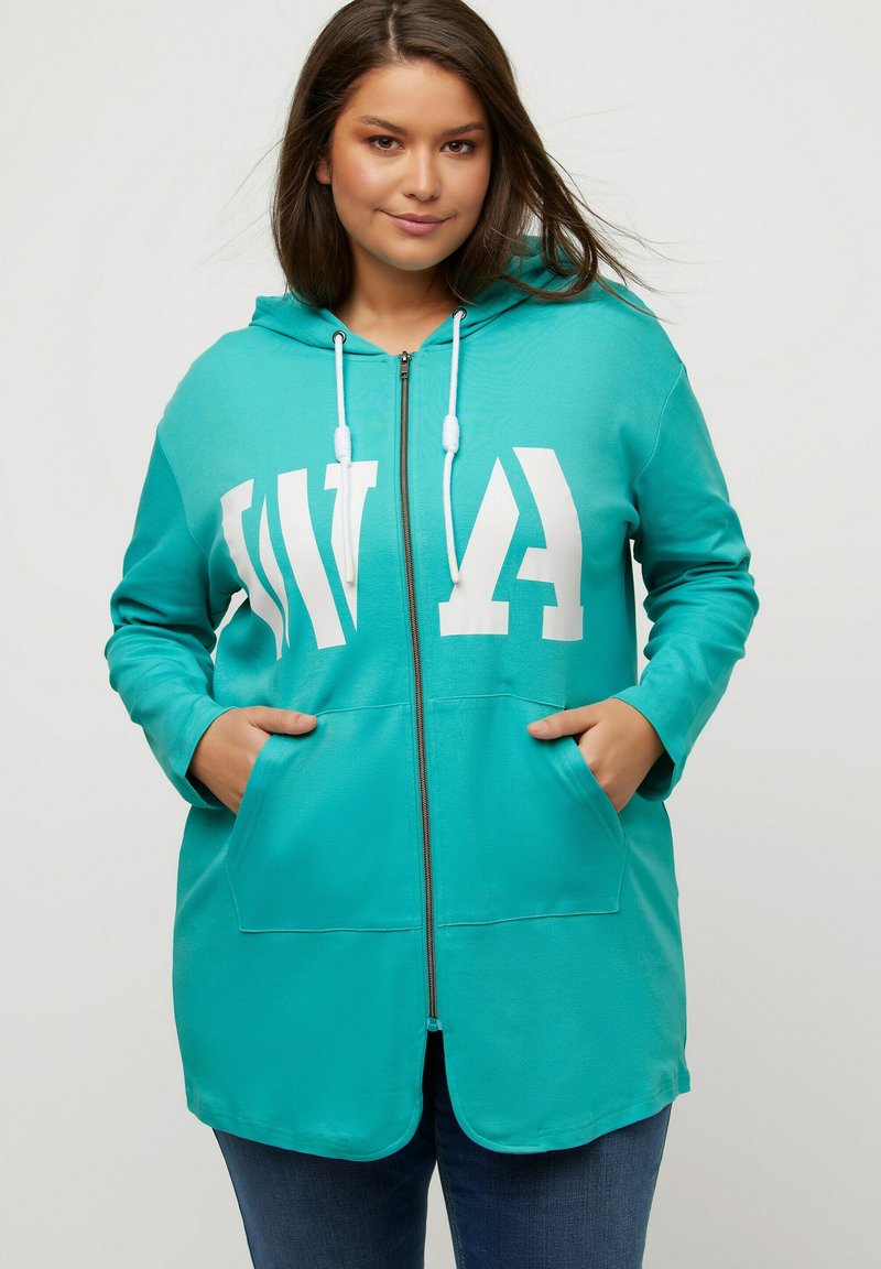 Chaqueta con capucha en color turquesa, con cremallera y texto en blanco, que presenta bolsillos frontales y una textura suave. Las mangas son largas y cuentan con lazos ajustables.