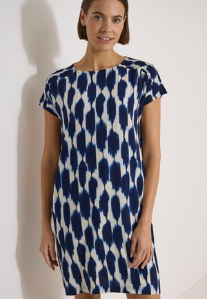 Femme aux cheveux bruns courts portant une robe à manches courtes avec un motif abstrait bleu et blanc, debout contre un mur clair.