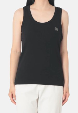 Donna che indossa una canotta nera senza maniche con un piccolo logo geometrico sul petto, abbinata a pantaloni bianchi.