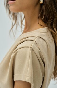 Top beige con detalles plisados en los hombros, tejido suave, mangas cortas y escote redondo, que muestra una sutil textura y elementos de diseño.