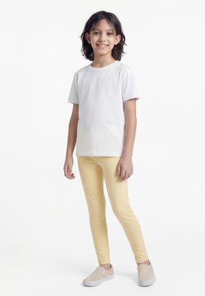 Kind met halflang donker haar staat glimlachend, draagt een effen wit T-shirt, lichtgele broek en beige instapschoenen.